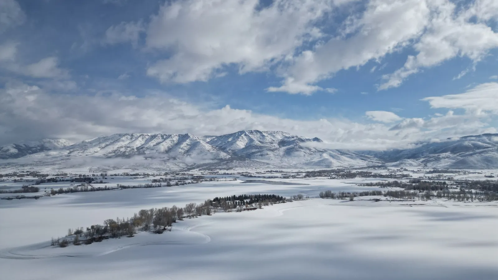 A snow-covered valley floor with scattered farmsteads and a frozen reservoir, ringed by the snowy peaks of the Ogden Valley under broken cloud